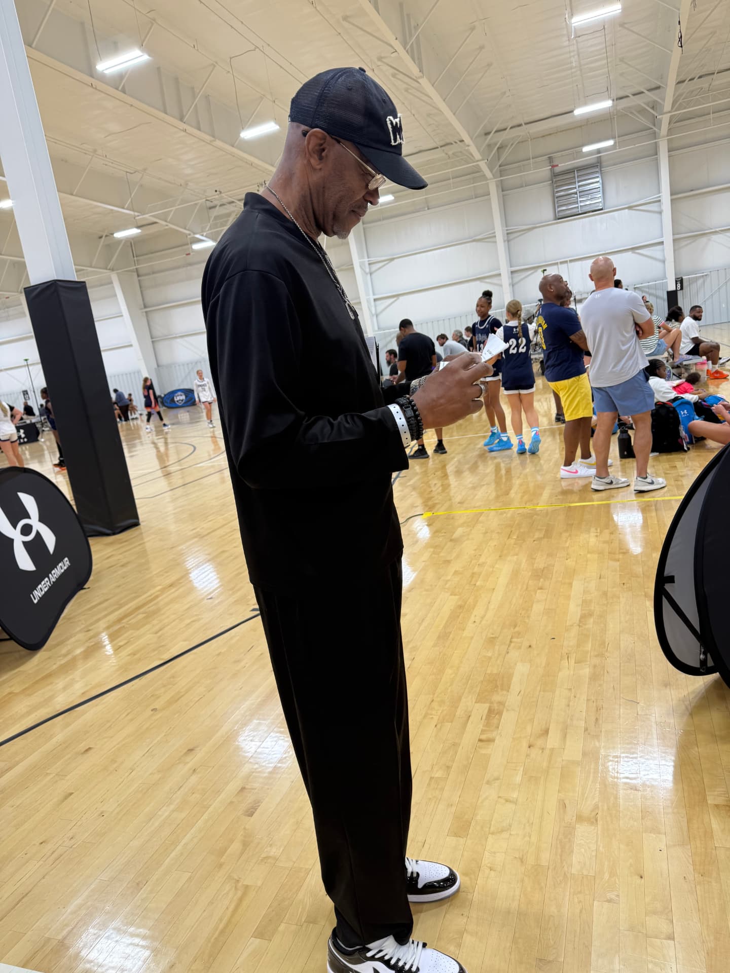 Side profile of Cary Noble in a gym, cap and glasses on, reviewing notes on his phone while players warm up on the court behind him.