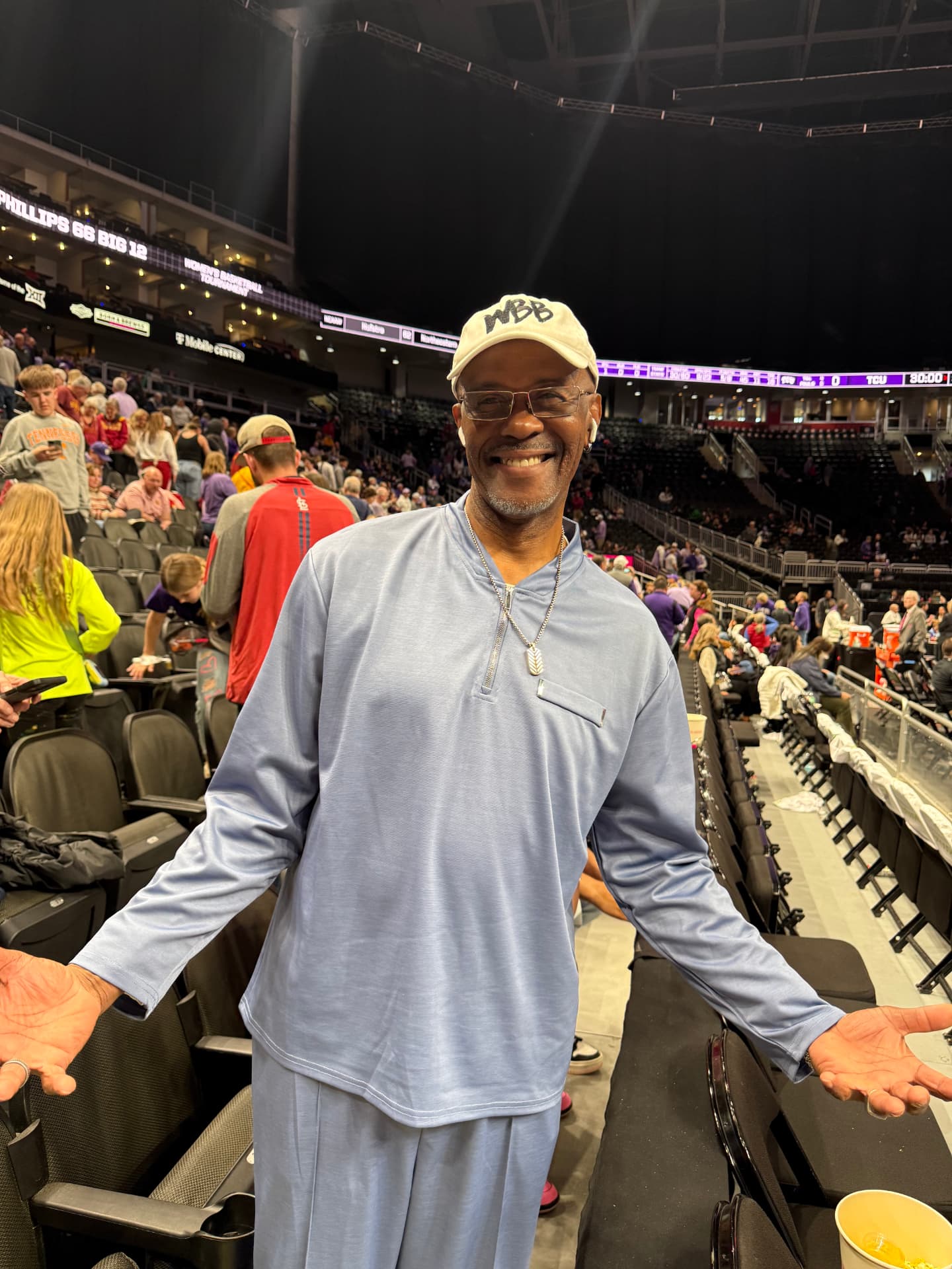 Cary Noble courtside at a Big 12 women's basketball tournament game, wearing a WBB cap and glasses with arms open in welcome.