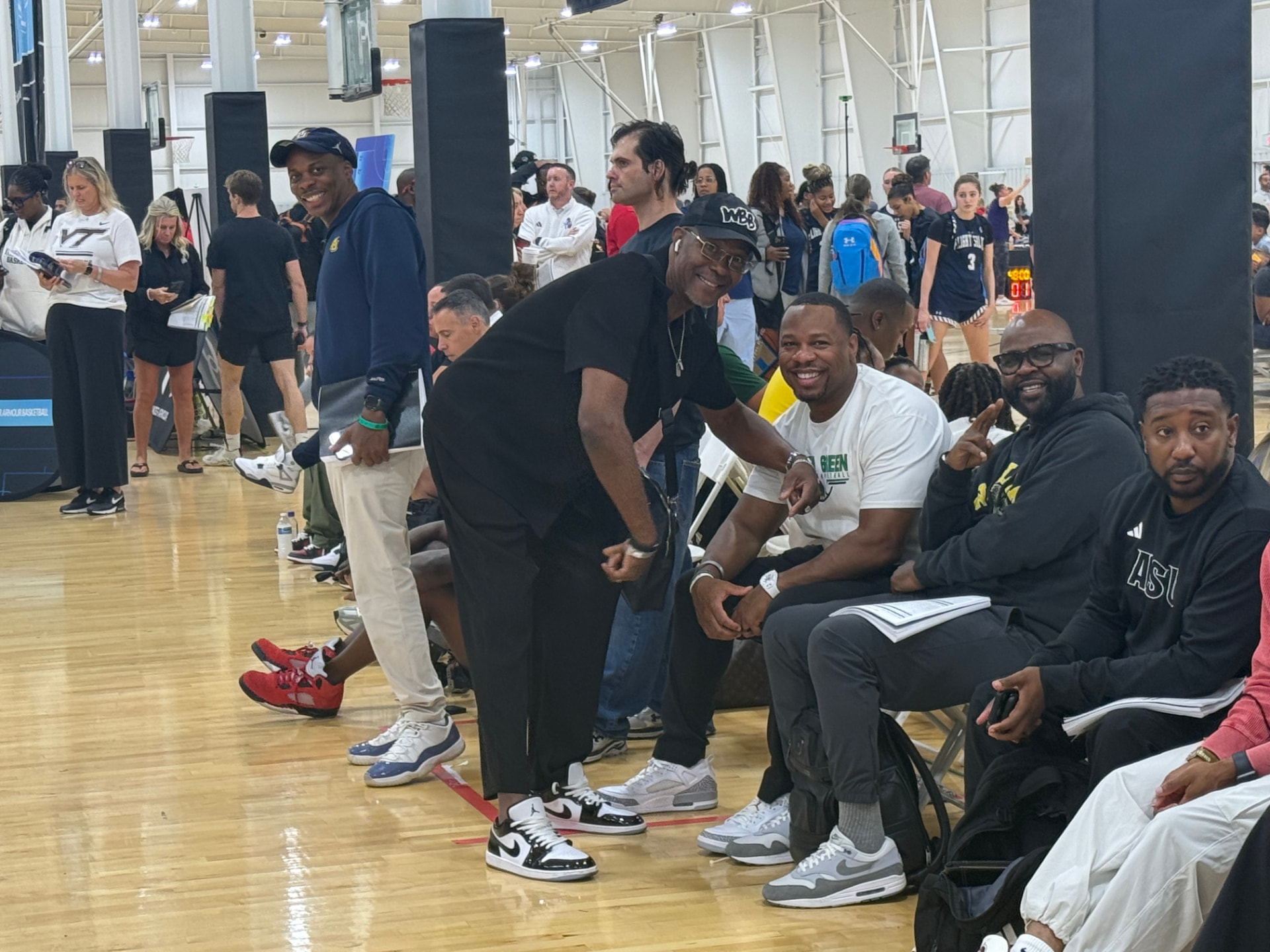 Cary Noble leaning in to talk with a row of seated college coaches on the sideline of a packed Ohio girls basketball event.